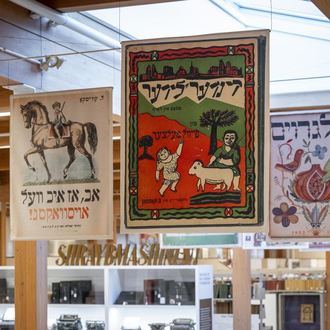Banners of Yiddish book covers hang in repository, glass case of typewriters in distance