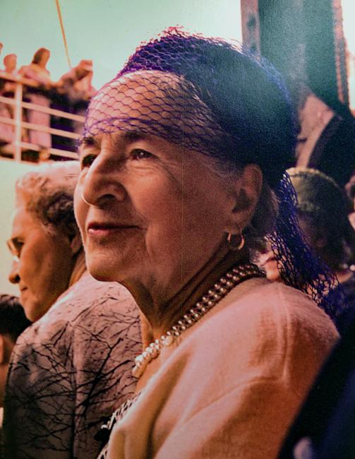 An older white woman wearing a purple lace headscarf and a pearl necklace fills the frame, with people in the background