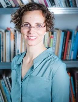 Woman with short brown hair and glasses smiling while posing in front of bookshelf. She is wearing a light blue dress shirt.