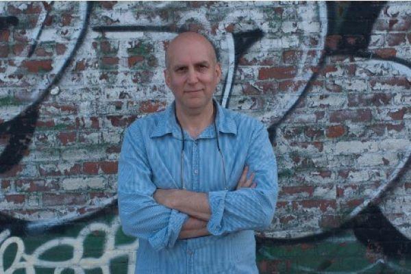 Man with blue dress shirt crossing his arms in front of graffitied wall.