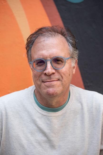 Man wearing glasses and white sweater in front of orange and black wall.